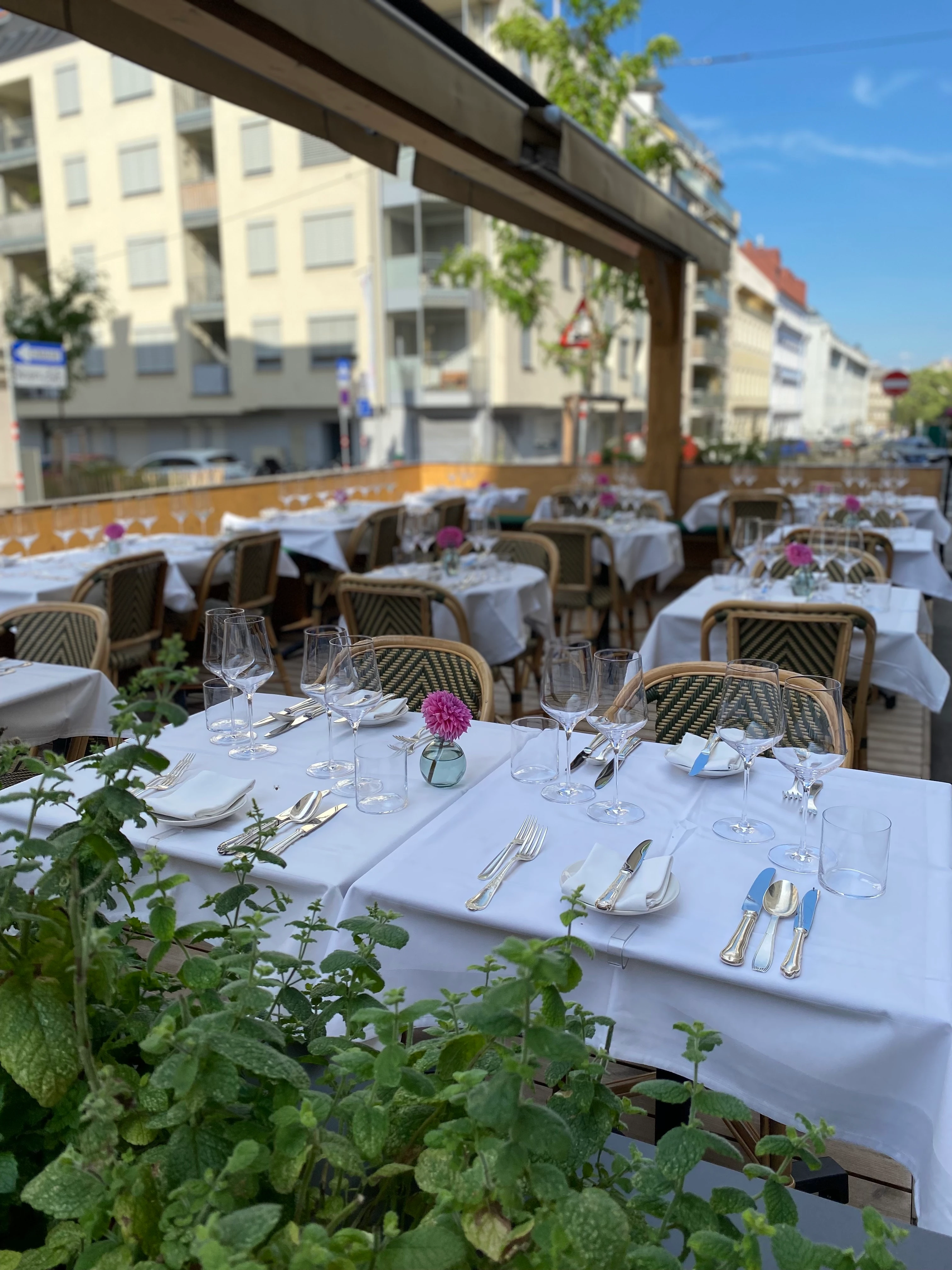 Elegantes Outdoor-Restaurant mit weißen Tischdecken, Besteck und Blumendekoration in einer städtischen Umgebung.